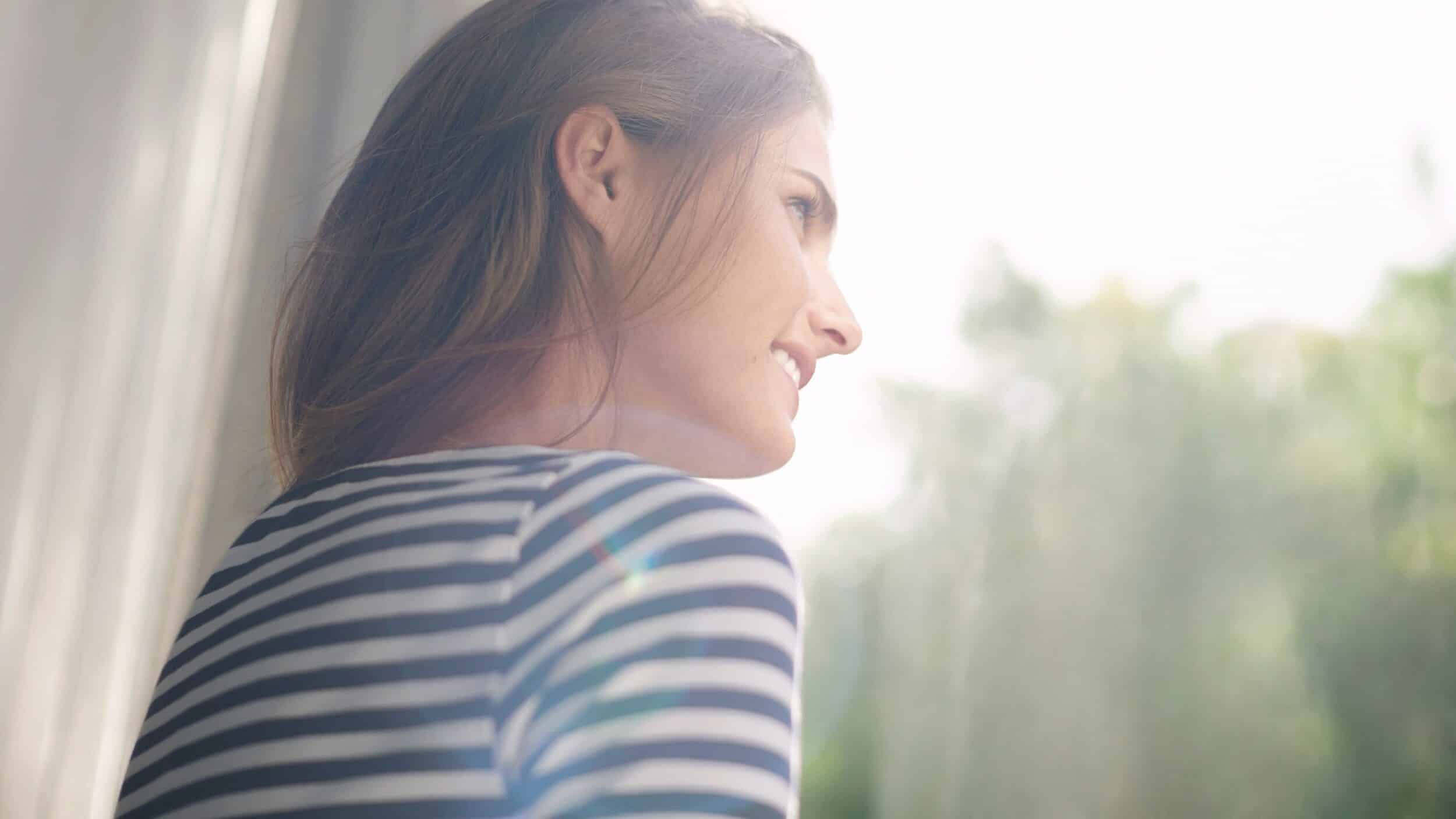 Une jeune femme regarde par la fenêtre et profite des rayons du soleil.