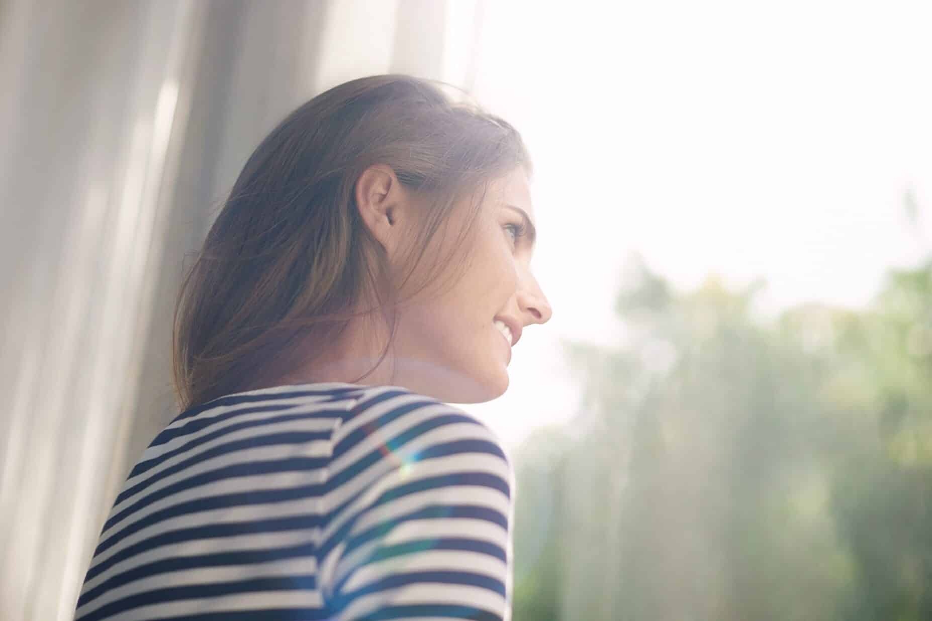 Une jeune femme regarde par la fenêtre et profite des rayons du soleil.