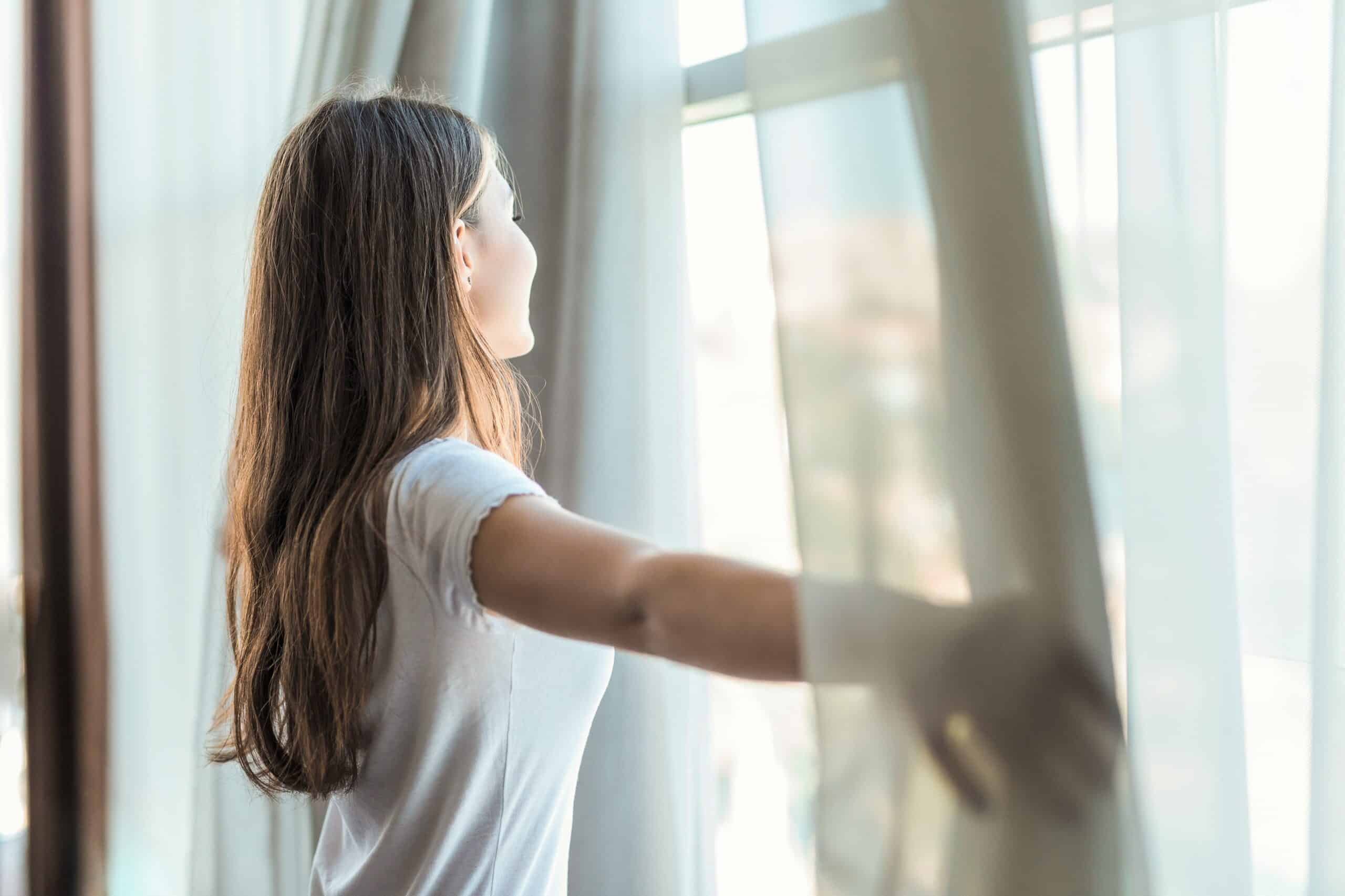 Frau steht am Fenster und schiebt Vorhänge zur Seite