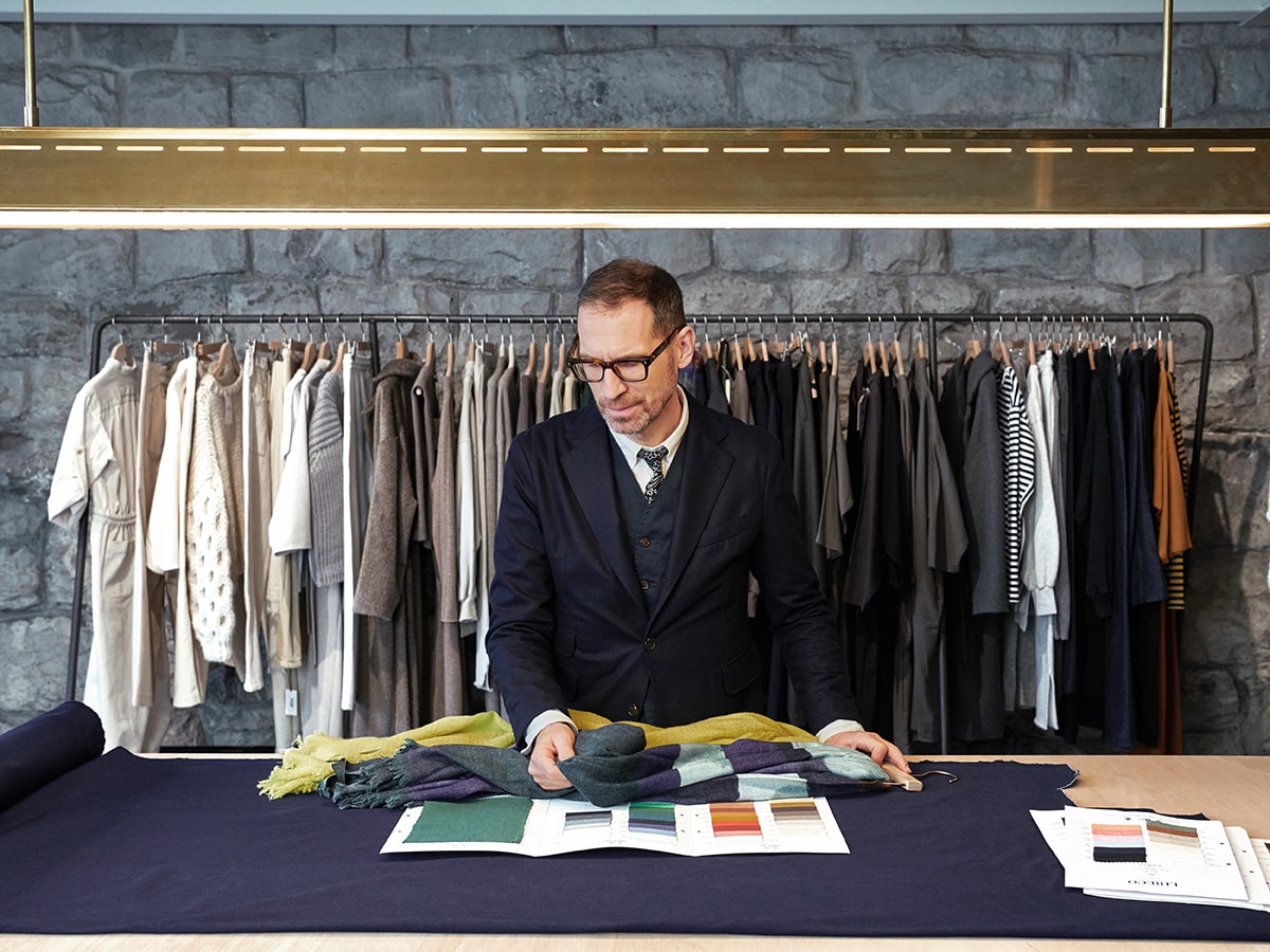 Meine Welt, meine Farben. Jeroen Van Rooijen bei der Arbeit bei der Auswahl eines Stoffes. Im Hintergrund Kleidungsstücke am Ständer.